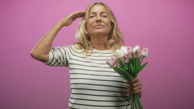 Woman in striped shirt holding pink tulips and gripping stems salutes with hand to forehead in studio; tribute serenity.