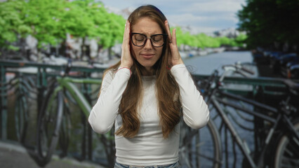 Fototapeta premium Woman holding hands to her ears and wincing on an amsterdam street bridge by canal; frustration stress.