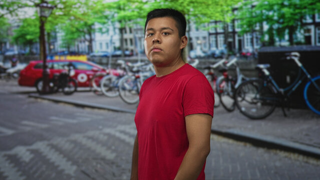 Man in a red t shirt standing and looking at camera on a city street, cobblestone pavement visible; quiet confidence.
