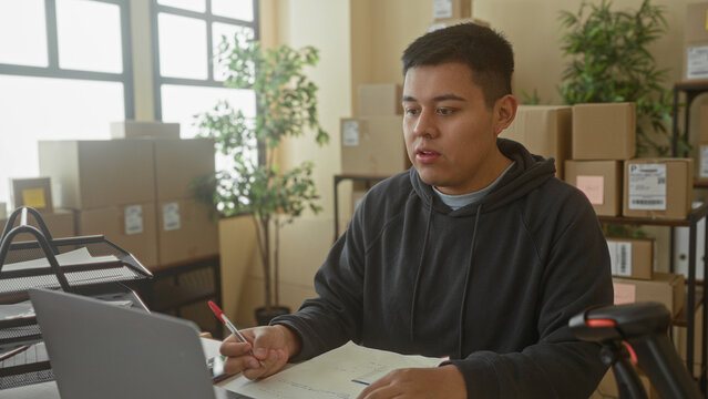 Man writing with pen at laptop surrounded by stacked shipping boxes and delivery labels in a building office; small business focus.