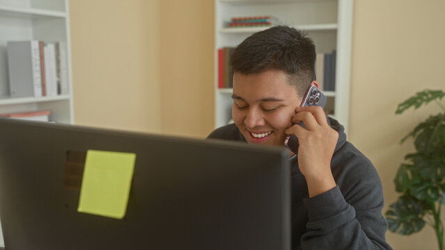 Man holds smartphone to ear, smiling at computer monitor while seated at a desk in a building; contentment.