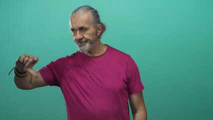 Fototapeta premium Man with gray hair and beard points finger and gives thumbs down in studio with teal backdrop and magenta tshirt; disapproval warning.