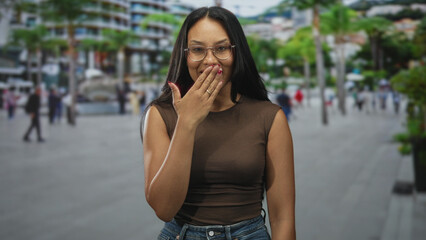 Fototapeta premium Young hispanic woman covers her mouth with hand while blowing a kiss on a busy urban street; playful affection.