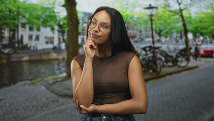 Woman wearing glasses crossing arms on street by canal with parked bicycles, lamp posts and cobblestone pavement  confidence reflection. © Krakenimages.com