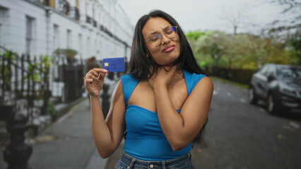 Woman holding creditcard with right hand while touching her neck and squinting, grimacing near...
