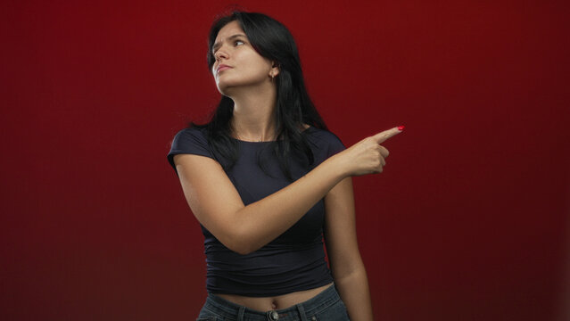 Young hispanic woman flicks finger dismissively in red studio with skeptical expression; defiance rejection.
