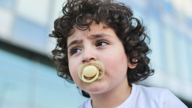 Young boy with curly hair using a pacifier outdoors looking up and away
