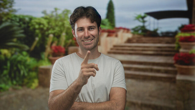 Man in white t shirt with crossed arms and visible forearms standing on stone building stairs among potted plants; quiet confidence.