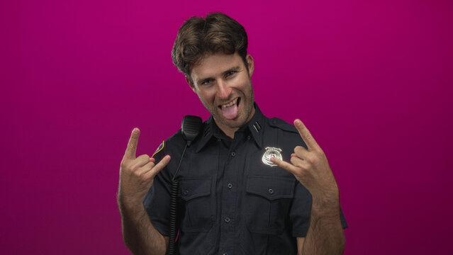 Young man policeman making rock horns with hands in studio against magenta backdrop; playful rebellion.