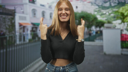 Obraz premium Redhead woman smiling with crossed fingers gesture on a street by white buildings and iron railings, wearing cropped top and jeans; hope luck.