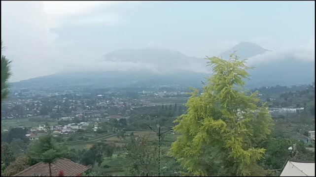 Beautiful and cloudy aerial view of the silhouettes of Mt. Gede and Mt. Pangrango shrouded in mist, overlooking rural agriculture fields and highland settlements in Puncak area, West Java, Indonesia
