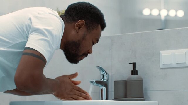 A man with dark skin is leaning over a sink while washing his face in a modern bathroom. The setting is bright, and there are toiletries on the counter.