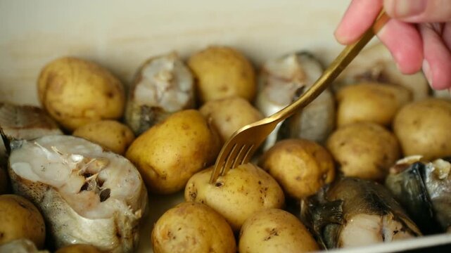 Golden fork piercing and lifting freshly baked potato from dish with oven cooked pollock fish steaks, demonstrating tenderness of the vegetables after being cooked in oven