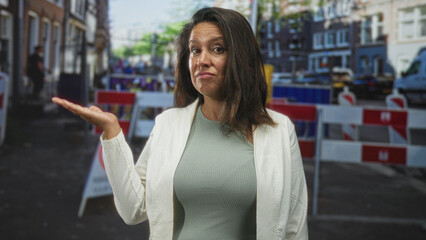 Fototapeta premium Woman shrugging with palm up hand at a blocked urban street with construction barriers and vans, looking unimpressed; frustration inconvenience.
