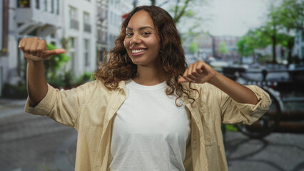 Woman thumbs pointing to chest with bright smile on a city street wearing white tshirt and beige shirt  confidence empowerment success. © Krakenimages.com
