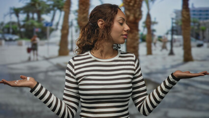 Young woman holding palms up in a shrug pose on a city street wearing a black and white striped shirt  thoughtful balance. © Krakenimages.com