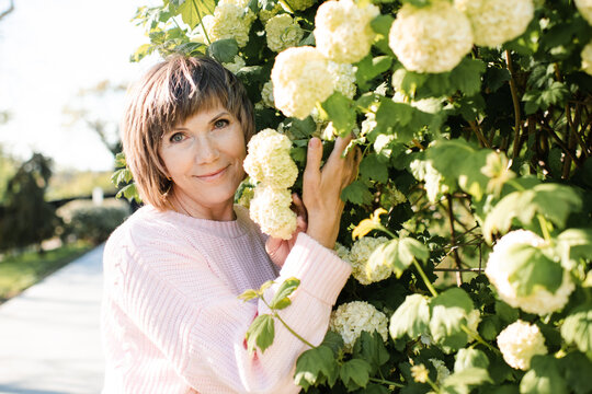 Blonde senior woman 55-60 year old posing with blooming bushes with white flowers in park outdoors. Mature wellbeing lifestyle. Springtime.