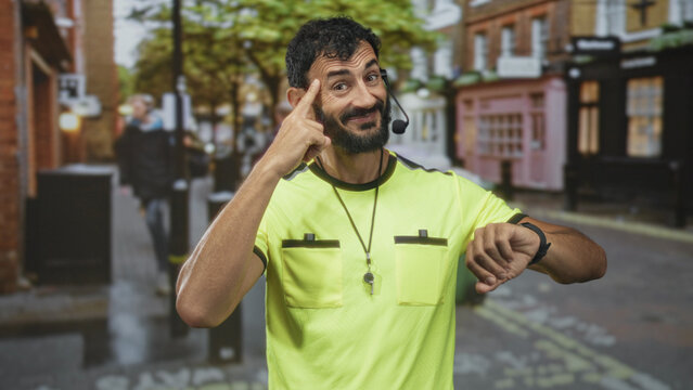 Man referee with whistle pointing to head and checking wristwatch while wearing headset microphone on urban street with shops and pavement; timekeeping urgency.