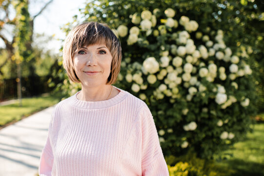 Blonde senior woman 55-60 year old posing with blooming bushes with white flowers in park outdoors. Mature wellbeing lifestyle. Springtime.