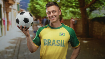 Fototapeta premium Man in brasil jersey holds soccer ball with right hand and points finger while smiling on street; joyful pride.
