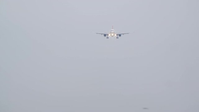 Commercial passenger airplane with landing lights on, making its final approach for landing in thick fog. The jet flies towards the camera in low visibility, hazy weather conditions.