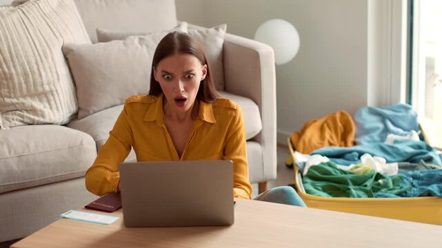 A woman with light skin sits on a couch looking shocked at her laptop. She holds a passport in her hand and has clothes scattered nearby. This scene takes place in a modern living room.