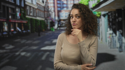 Obraz premium Woman with hand on chin at a city street crosswalk, neutral expression and folded arm across body, visible ring on finger; quiet contemplation.