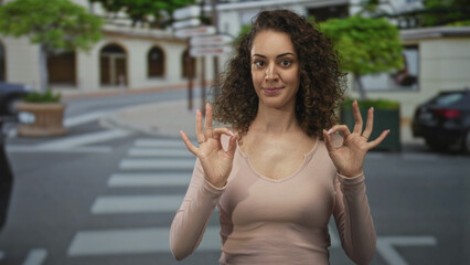 Fototapeta premium Young hispanic woman making ok sign with both hands while standing on a city crosswalk and sidewalk near parked cars and planters; confidence.
