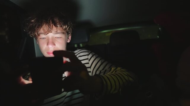 A young man with curly hair is deeply focused on his smartphone screen in the dark back seat of a car at night. The phone's bright screen illuminates his face as he scrolls or interacts,