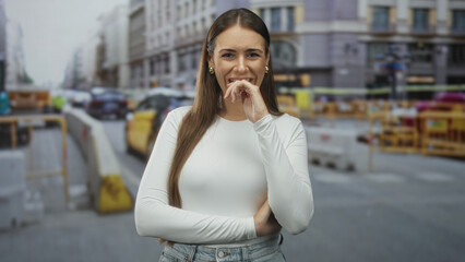 Fototapeta premium Young woman in white long sleeve top, hand near mouth in a shy laugh and crossed arms, standing on a busy city street with yellow taxis and construction barriers visible; playful confidence.