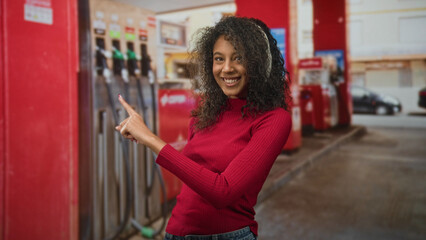 Young woman with curly hair wearing white headphones points finger toward a gas pump at a street station while smiling in a red sweater and denim jeans  joyful curiosity. © Krakenimages.com