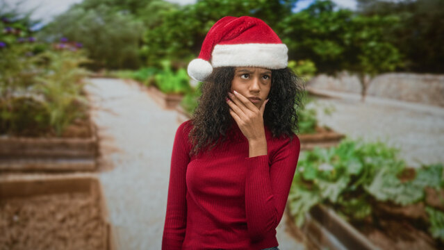 Woman in santa hat touches chin with hand, curly hair visible, red sweater, hand to chin gesture on a street garden path; thoughtful holiday introspection.