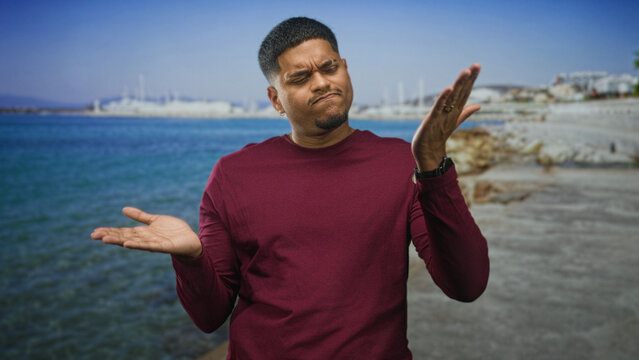 Hispanic man with beard touching chin and folding arm on a street by a marina and rocky shore; contemplation.