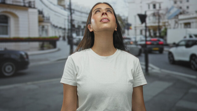 Young woman wearing white tshirt looks up at the sky on a busy city street lined with cars and buildings; optimism.