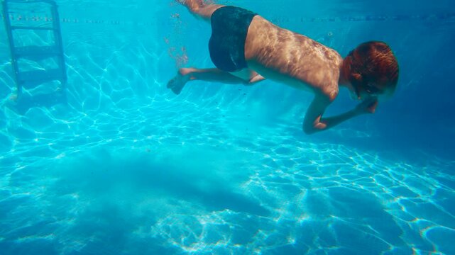 Happy ten year old boy having fun in a swimming pool, diving underwater and spinning in water. Active childhood leisure and summer recreation. Slowmotion video.