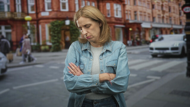 Woman in denim jacket folding arms and looking down at pavement by a zebra crossing on a busy urban street with brick storefronts and passing car; sadness reflection.