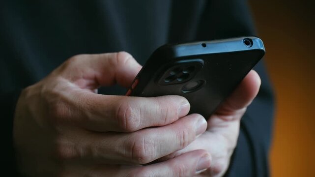 Close up view of male hands holding modern smartphone, with thumbs actively typing message, browsing social media, or scrolling through news feed in a well lit indoor setting. Person texting message