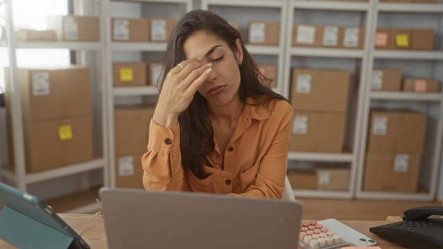 Woman pinches nose at laptop and tablet amid stacked parcel boxes on a delivery office desk in a building; frustration fatigue.