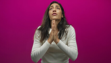 Obraz premium Young hispanic woman praying with clasped hands in studio against magenta backdrop; hopeful plea for comfort.