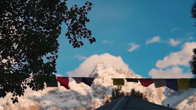 Prayer flags waving in the wind in front of snowy Himalayan mountain peak during the sunset as seen from Tayul monastery near Keylong in Lahaul Valley, Himachal Pradesh, India. Scenic natural view.