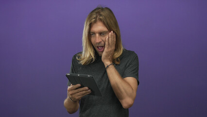 Fototapeta premium Blond man with long hair in dark shirt points finger to tablet with mouth open in purple studio; surprise.