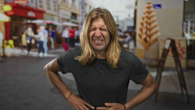 Blond man with long hair visibly grimaces as he clutches stomach on street; pain discomfort nausea distress.