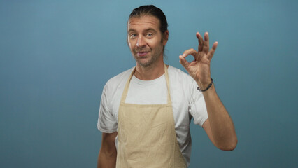Man with long blond hair wearing beige apron over white shirt making ok sign gesture with bare...