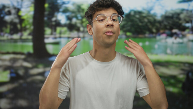 Young hispanic man wearing white t shirt and glasses gesturing hands while speaking beside a green pond in a park with trees; enthusiasm.