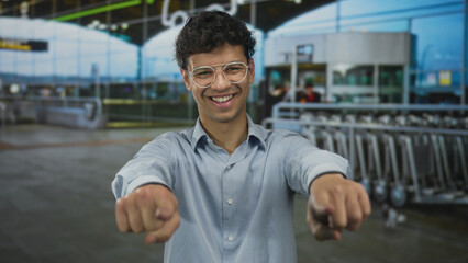 Fototapeta premium Man pointing fingers at camera in busy airport terminal with glass windows and luggage carts behind him; confidence.