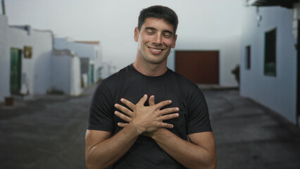 Fototapeta premium Smiling hispanic man wearing black shirt holds hands to chest on historic stone street in old town; self love gratitude.