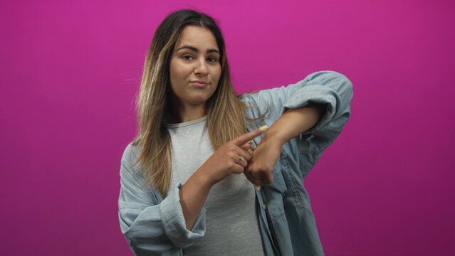 Hispanic woman pointing finger to wrist and tapping wrist in pink studio wearing denim shirt and gray tee, long hair visible; impatience.