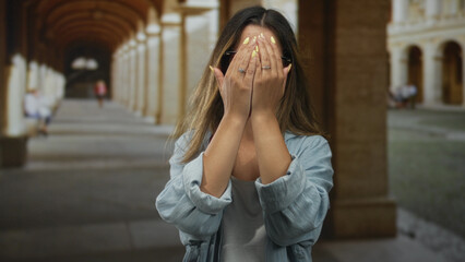 Fototapeta premium Woman with hands over face on old town street, squinting, showing yellow nails and rings while wearing glasses and denim jacket; frustration privacy.