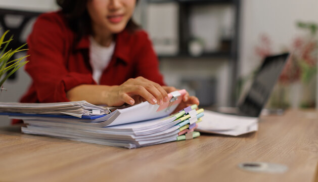Woman organizing thick stack of office documents and reports with colorful binder clips on a desk in a modern corporate workspace, hands sorting paperwork and files