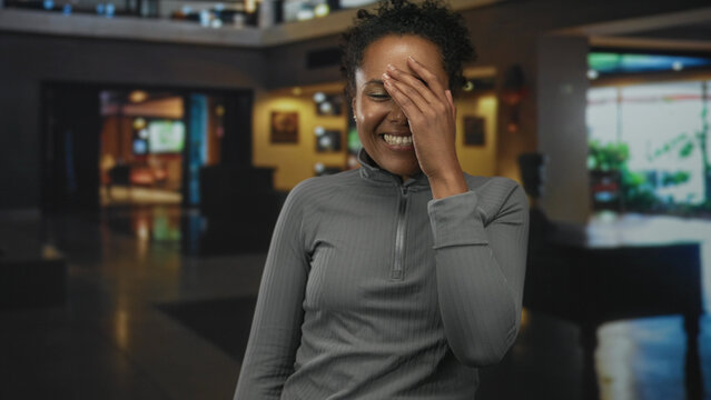 Young african american woman laughing and covering her face with her hand in a hotel lobby near seating area and reception desk; bashful happiness.
